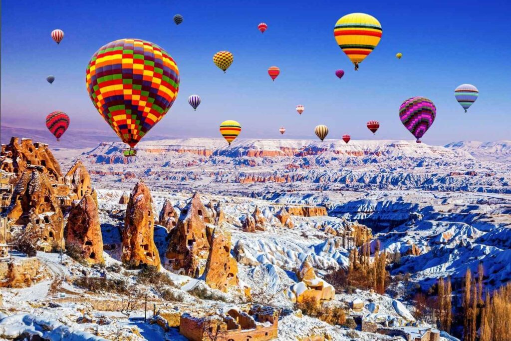 Vibrant hot air balloons soar above the snow-dusted rock formations of Cappadocia, Turkey, on a clear winter day.