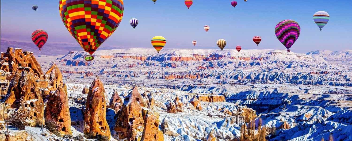 Vibrant hot air balloons soar above the snow-dusted rock formations of Cappadocia, Turkey, on a clear winter day.