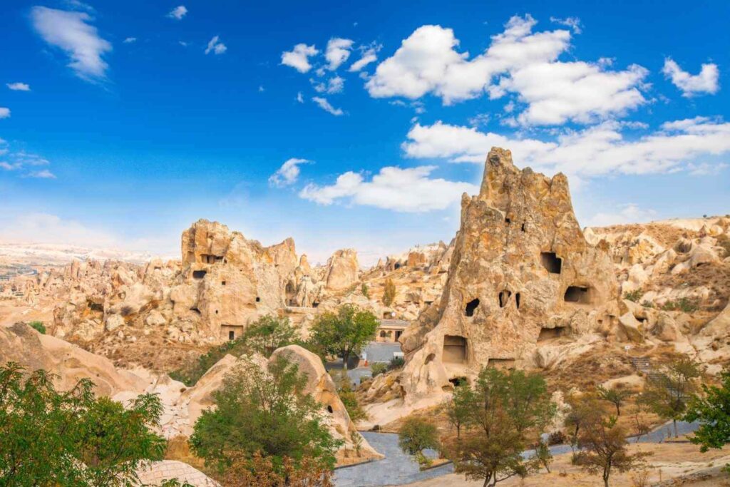 Rock formations with ancient cave dwellings in Cappadocia, Turkey, set against a bright blue sky with scattered clouds.