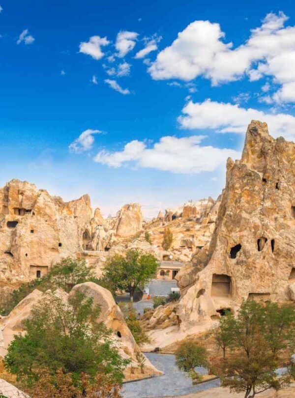 Rock formations with ancient cave dwellings in Cappadocia, Turkey, set against a bright blue sky with scattered clouds.