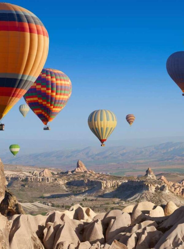 Vibrant hot air balloons floating over the rocky landscape of Cappadocia, Turkey, under a clear blue sky.