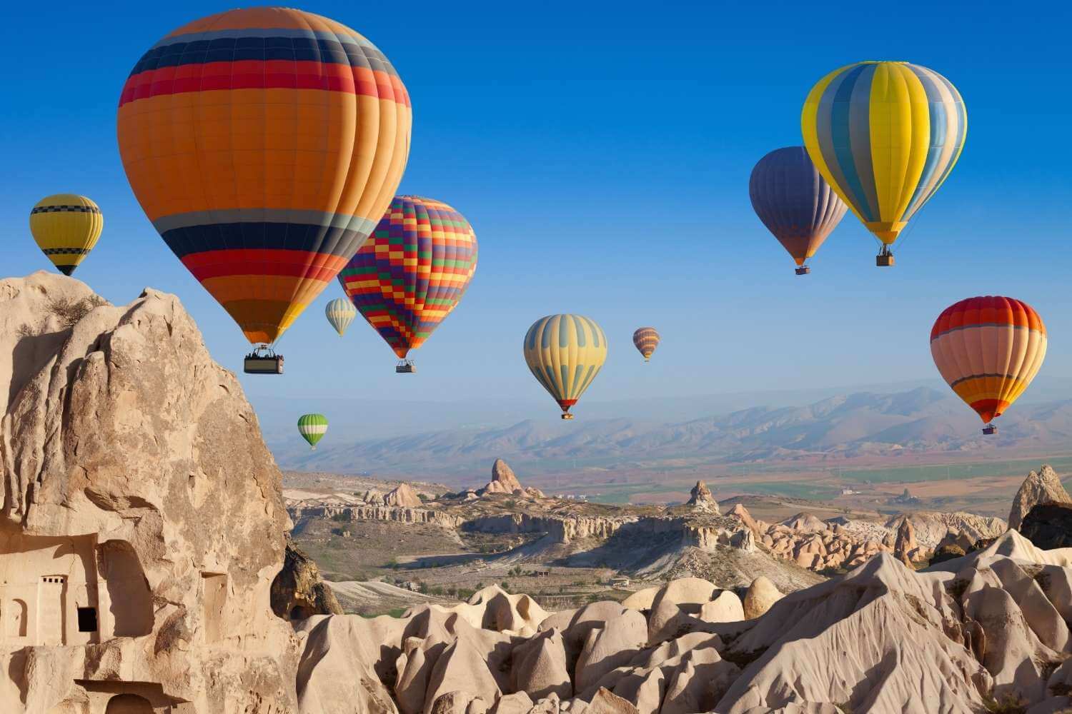 Vibrant hot air balloons floating over the rocky landscape of Cappadocia, Turkey, under a clear blue sky.