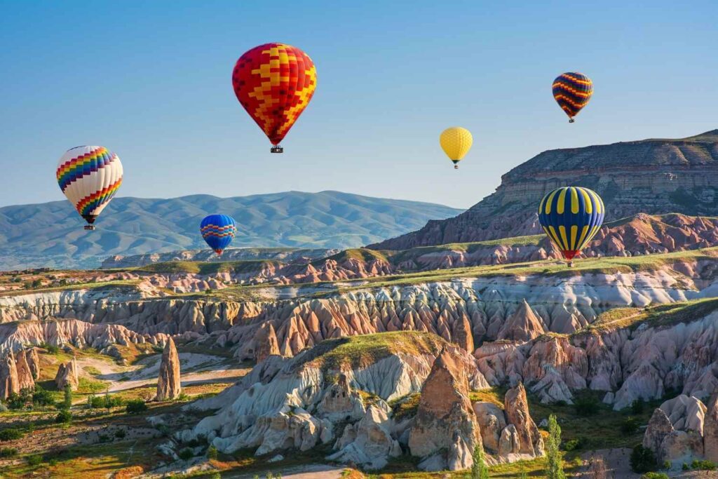 Colorful hot air balloons drift over Cappadocia's rugged landscape with rocky formations and distant mountains.