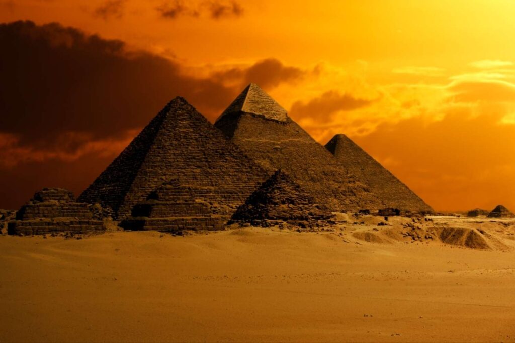 The Pyramids of Giza at sunset, silhouetted against a glowing orange sky with scattered clouds, surrounded by desert sands.