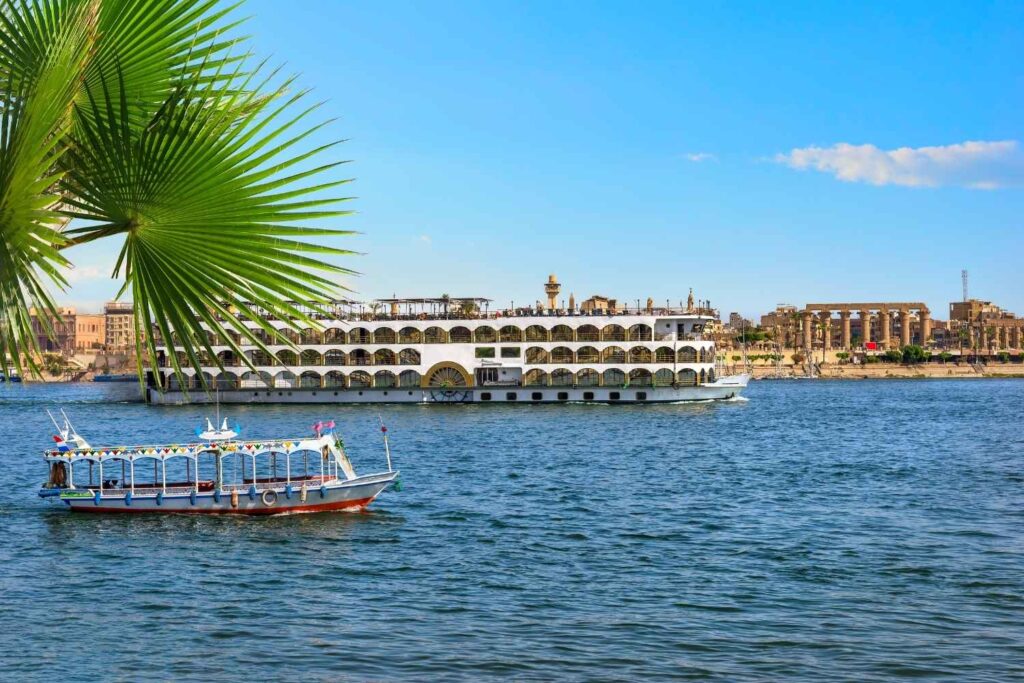 A scenic Nile River cruise with a traditional boat in the foreground and a luxury cruise ship passing near ancient Egyptian temples in the background.