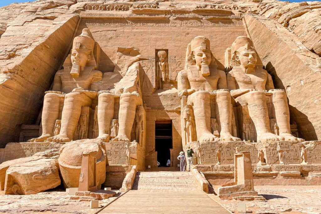 The Great Temple of Abu Simbel in Egypt, featuring colossal statues of Ramses II carved into a sandstone cliff, with intricate carvings and tourists nearby.