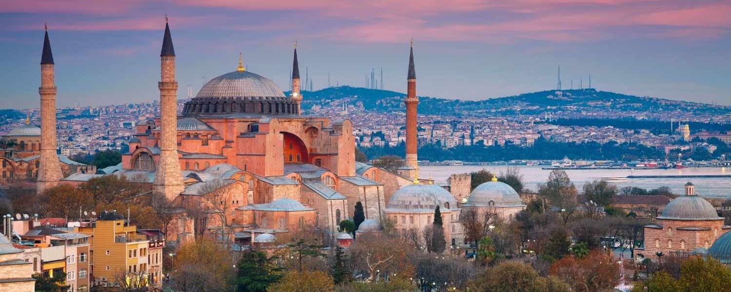 Hagia Sophia in Istanbul at sunset, featuring its domes and minarets with a cityscape and water in the background.