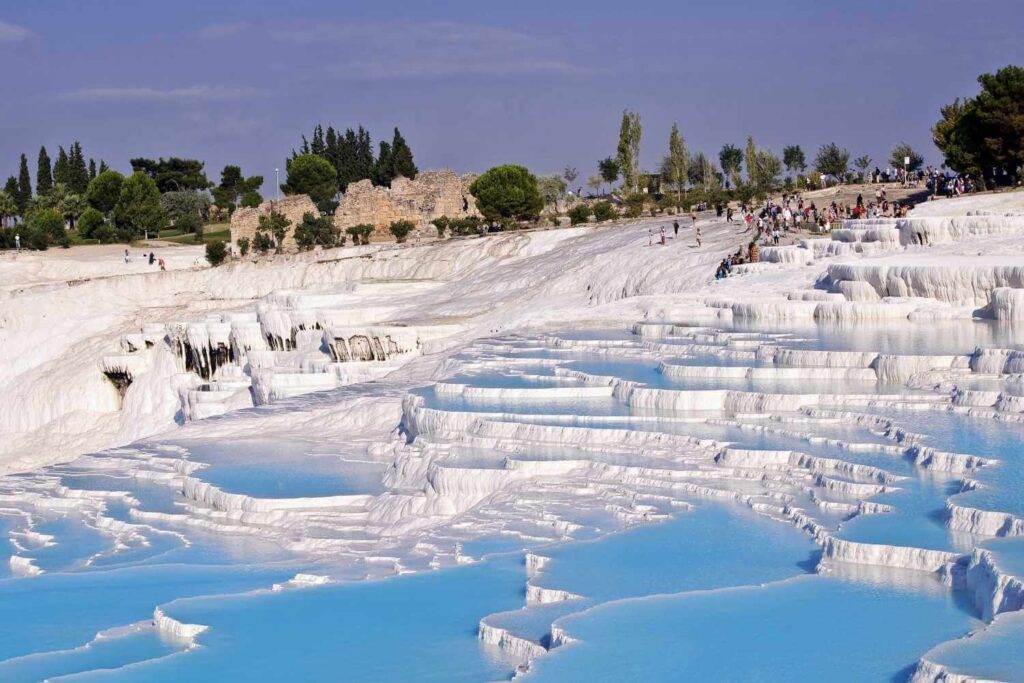 Tourists walking along the white travertine terraces filled with blue thermal water at Pamukkale, Turkey, on a sunny day.