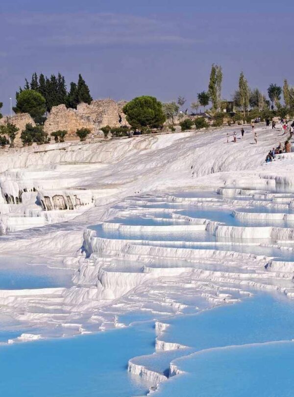 Tourists walking along the white travertine terraces filled with blue thermal water at Pamukkale, Turkey, on a sunny day.