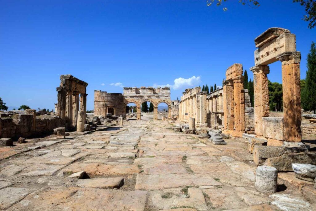 The ancient stone-paved main street of Hierapolis, Turkey, lined with ruins and arches under a bright blue sky.