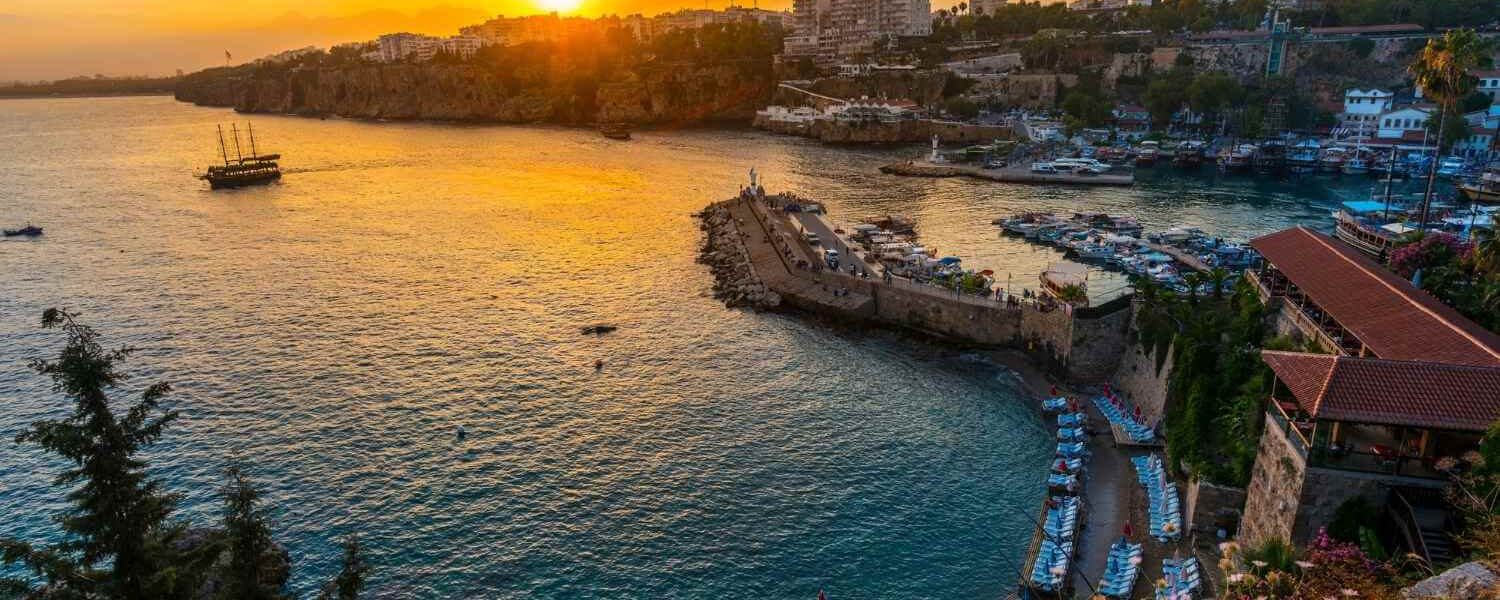 Coastal view of Antalya, Turkey at sunset, featuring a curved shoreline, historic harbor, and cityscape in the background.