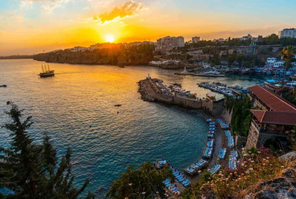 Coastal view of Antalya, Turkey at sunset, featuring a curved shoreline, historic harbor, and cityscape in the background.