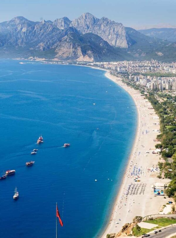 Aerial view of Antalya's Konyaaltı Beach, Turkey, with clear blue water, sandy shoreline, and majestic mountains in the background.