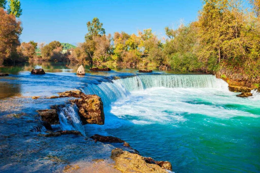 Scenic view of Manavgat Waterfall in Turkey, with cascading water over wide rocks surrounded by lush greenery.