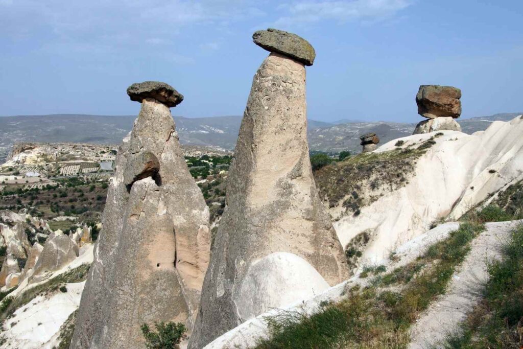 Tall, cone-shaped rock formations known as fairy chimneys in Cappadocia, topped with large stone caps against a mountainous backdrop.