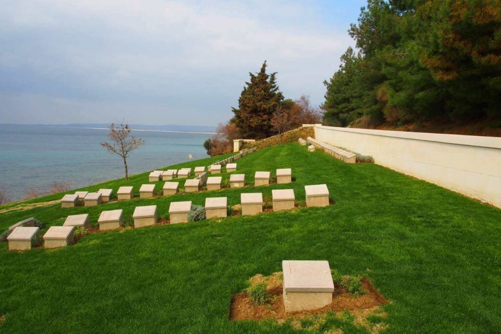 Military cemetery on a grassy slope overlooking the sea, with rows of white headstones and surrounding trees.