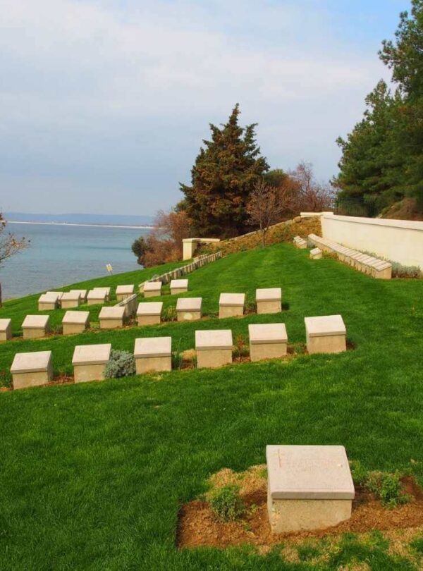 Military cemetery on a grassy slope overlooking the sea, with rows of white headstones and surrounding trees.