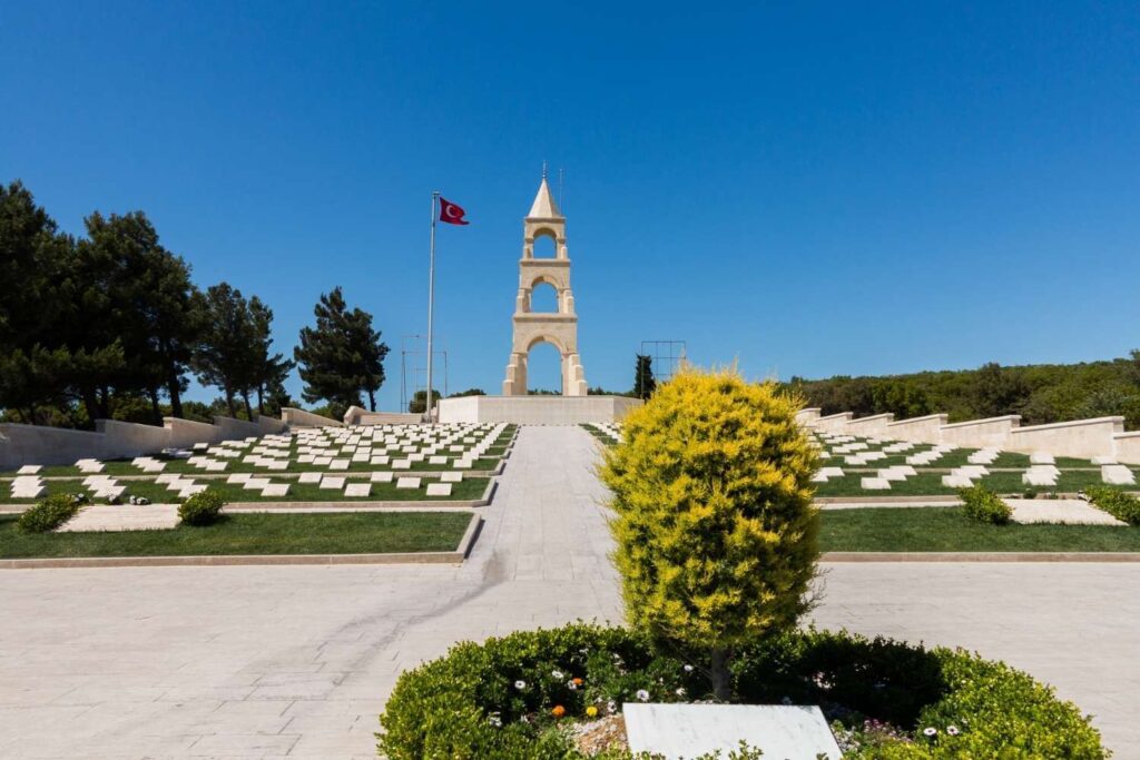 Memorial cemetery with rows of headstones, a central monument, and a Turkish flag, under a clear blue sky.