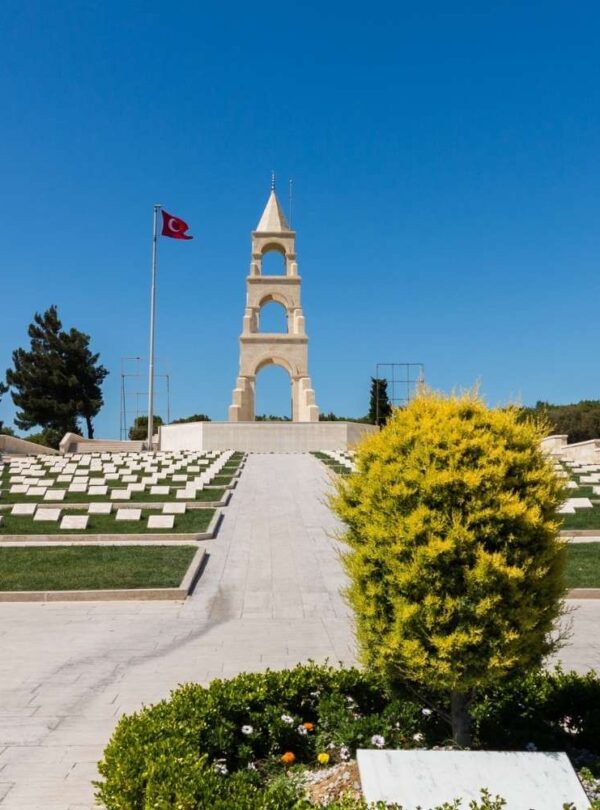 Memorial cemetery with rows of headstones, a central monument, and a Turkish flag, under a clear blue sky.