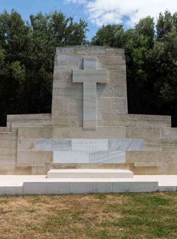 Stone memorial with a cross and engraved inscription, surrounded by greenery, honoring fallen soldiers.