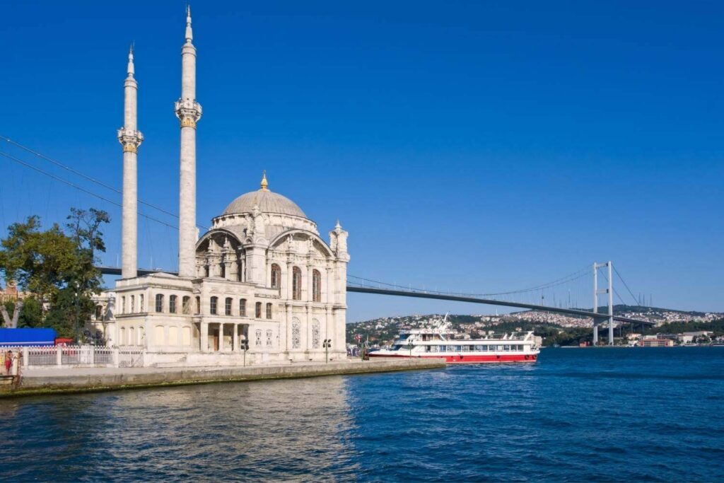 Ortaköy Mosque with twin minarets on the Bosphorus waterfront, with a ferry and the Bosphorus Bridge under a clear blue sky.