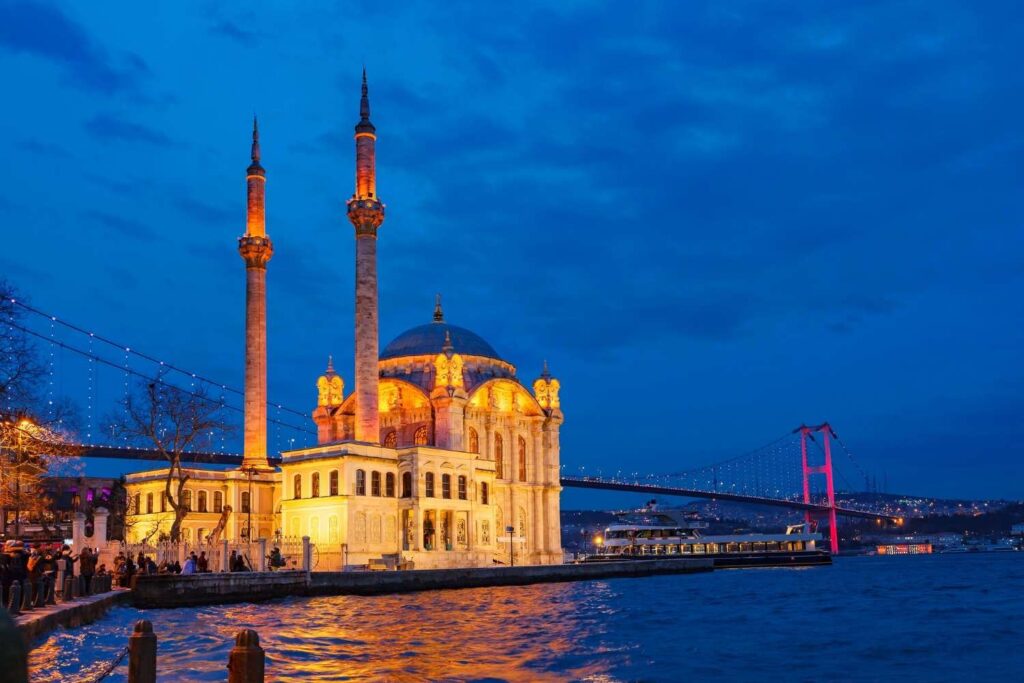 Ortaköy Mosque illuminated in warm lights at dusk, with the Bosphorus Bridge lit in red, reflecting on the water in Istanbul.