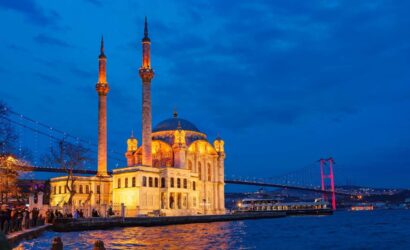 Ortaköy Mosque illuminated in warm lights at dusk, with the Bosphorus Bridge lit in red, reflecting on the water in Istanbul.