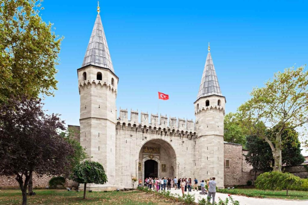 Entrance gate of Topkapi Palace in Istanbul, with tourists and Turkish flag under a clear blue sky.