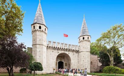 Entrance gate of Topkapi Palace in Istanbul, with tourists and Turkish flag under a clear blue sky.