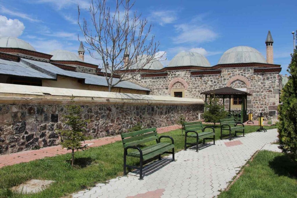 xterior of the Sultan Selim Mosque in Konya, Turkey, featuring stone walls, domed roofs, and a garden path with benches.