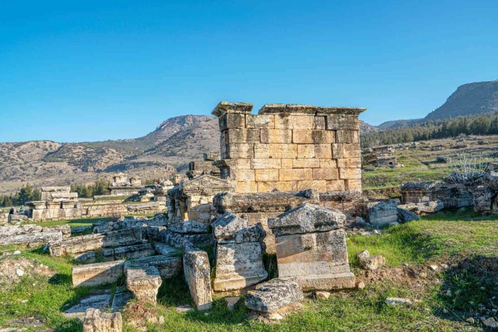 Stone ruins of an ancient structure with scattered blocks on a grassy field, set against a backdrop of mountains and a clear blue sky.