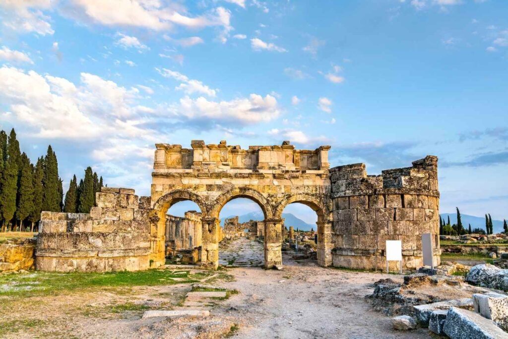 Historic stone gate of Hierapolis with three arched entrances, set against a backdrop of trees and blue sky in Turkey.