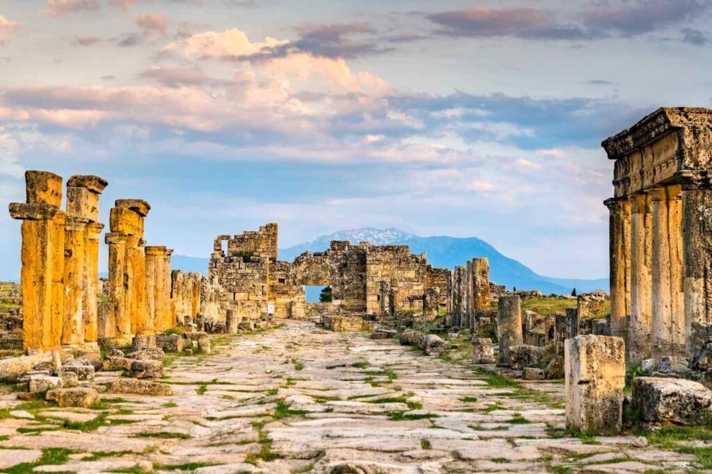 Stone-paved street of the ancient city of Hierapolis, Turkey, lined with weathered columns and ruins against a mountainous backdrop.