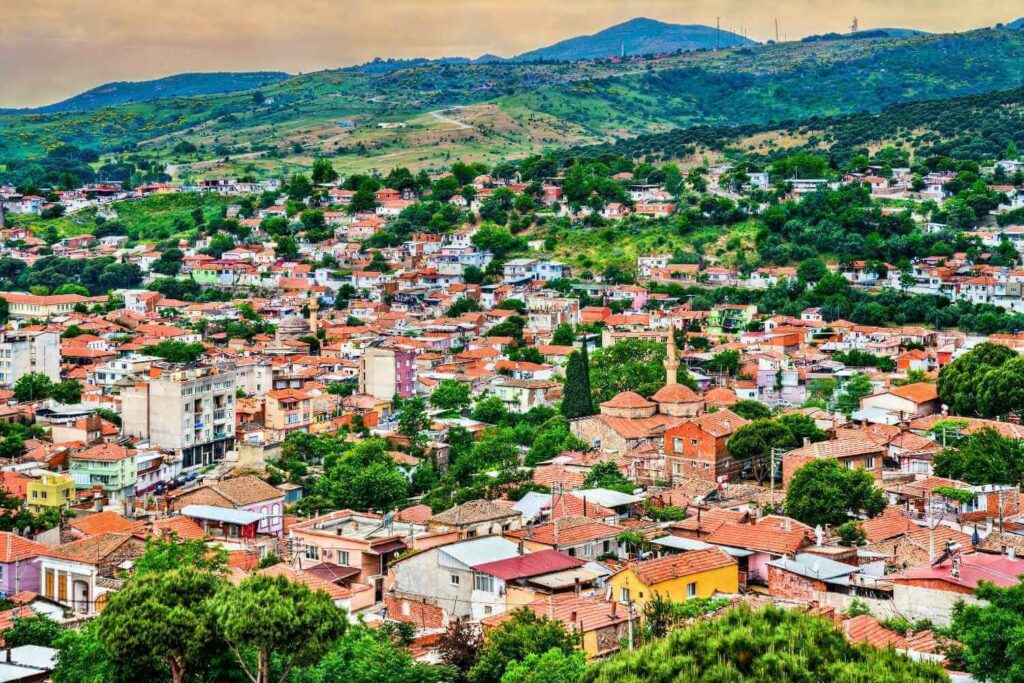 Aerial view of a Turkish town with red-roofed houses and a mosque, surrounded by lush green hills under a cloudy sky.