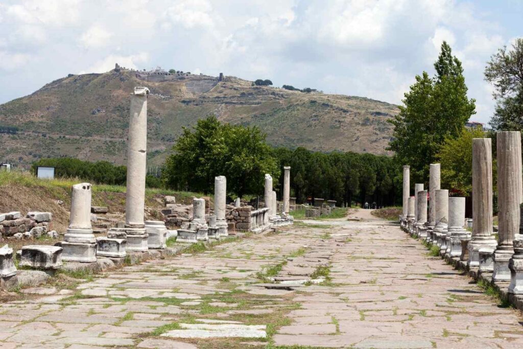 Ancient stone pathway lined with marble columns in the ruins of Pergamon, Turkey, with a hill in the background.