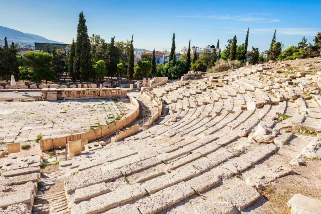 Stone seating of the ancient Theater of Dionysus in Athens, Greece, surrounded by trees and cityscape under a clear sky.