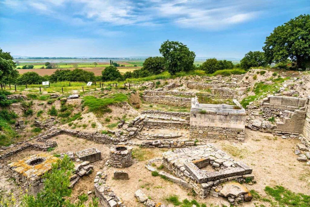 Ruins of ancient Troy featuring stone walls and foundations, set against a scenic landscape and cloudy sky.