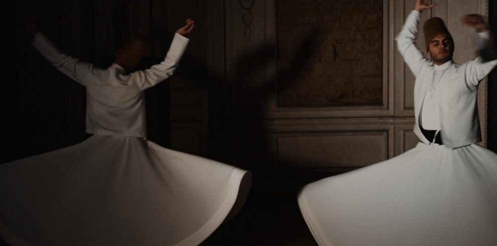 Two whirling dervishes in white robes perform a spiritual dance in a dimly lit room, showcasing traditional Sufi culture.