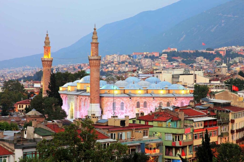 Bursa Grand Mosque with illuminated twin minarets and domed roof, surrounded by colorful city buildings and mountains at dusk.