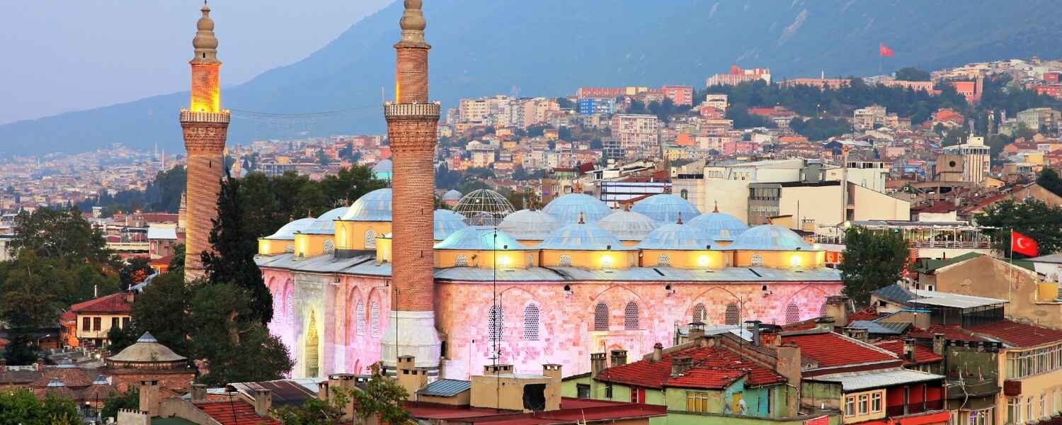 Bursa Grand Mosque with illuminated twin minarets and domed roof, surrounded by colorful city buildings and mountains at dusk.