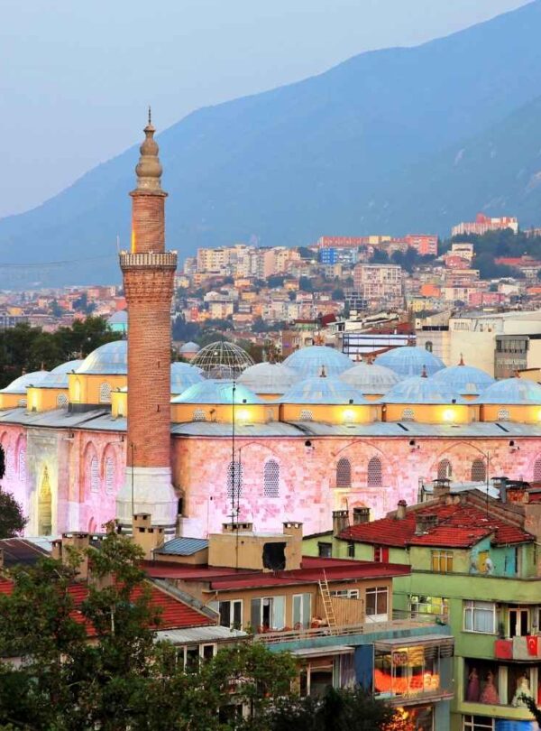 Bursa Grand Mosque with illuminated twin minarets and domed roof, surrounded by colorful city buildings and mountains at dusk.